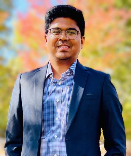A young man in a suit jacket smiles at the camera against a background of fall trees
