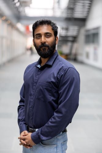 A young man standing in an academic hallway smiles at the camera.