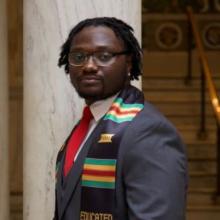 An African American young man with glasses, suit, tie, and honors scarf looks towards the camera